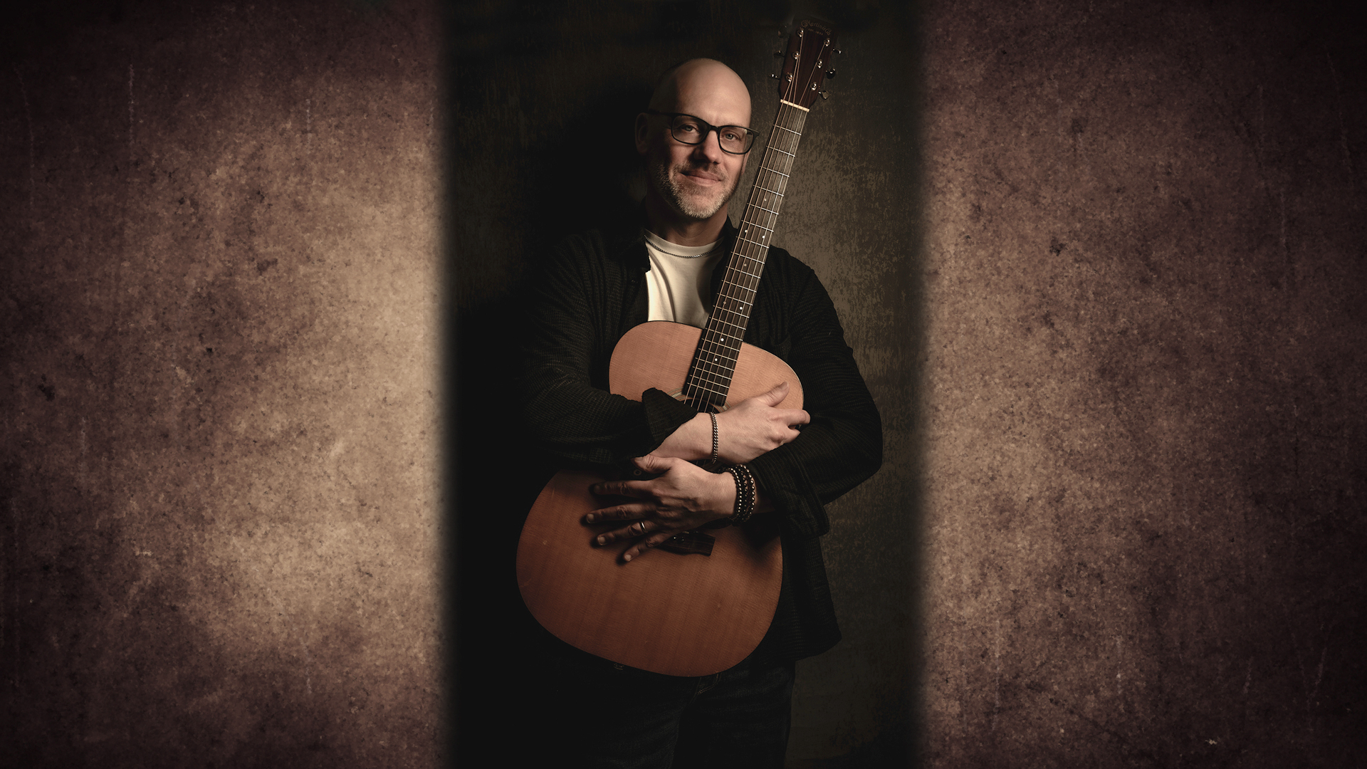 Fingerstyle guitarist Ben Woolman stands smiling with his arms wrapped around his acoustic guitar in dark, moody lighting.