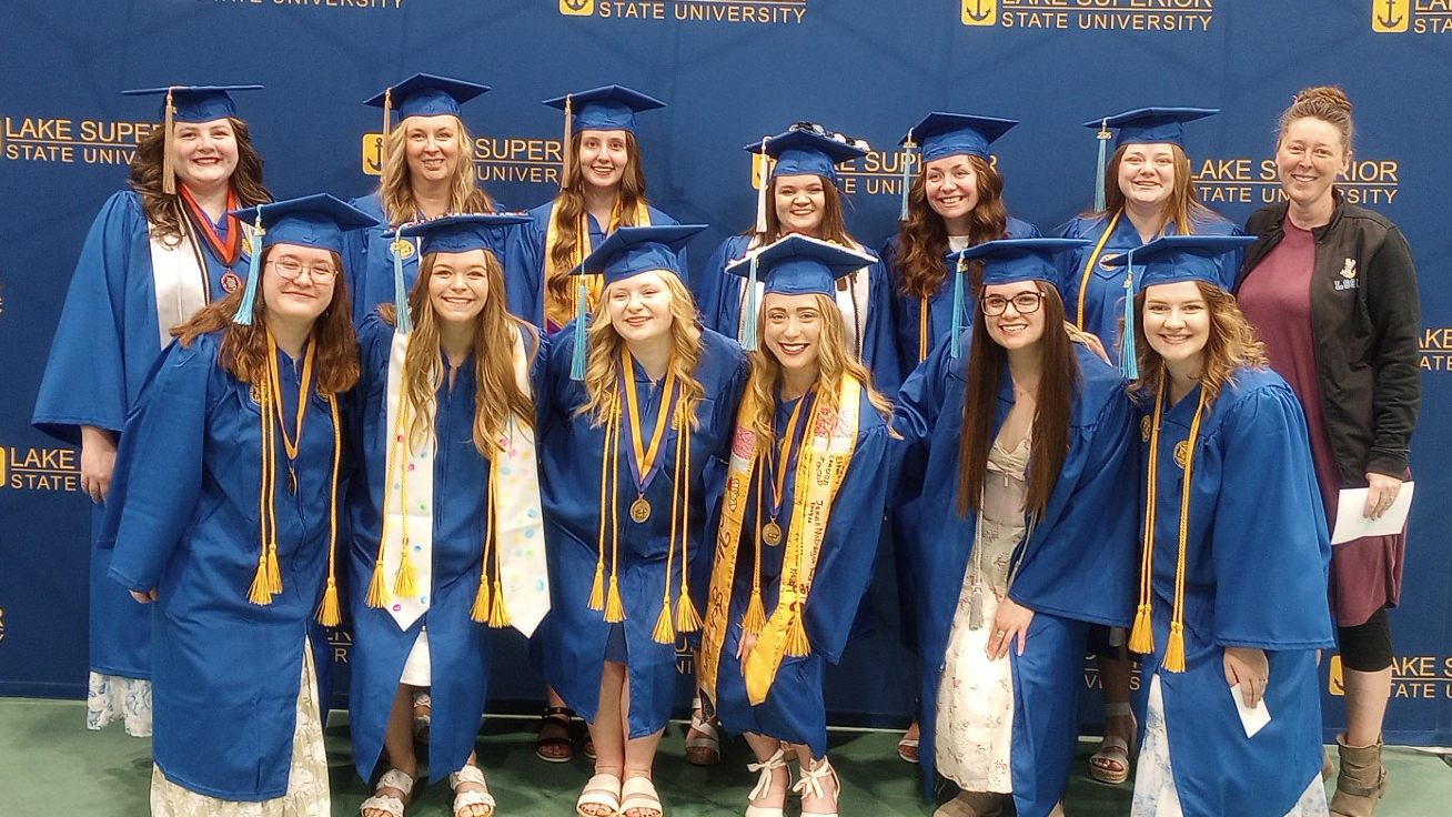 A dozen happy grads in caps and gowns smile in front of a Lake Superior State University logo backdrop with Director of LSSU Escanaba/Iron Mountain & Petoskey Regional Centers Heidi Rife.