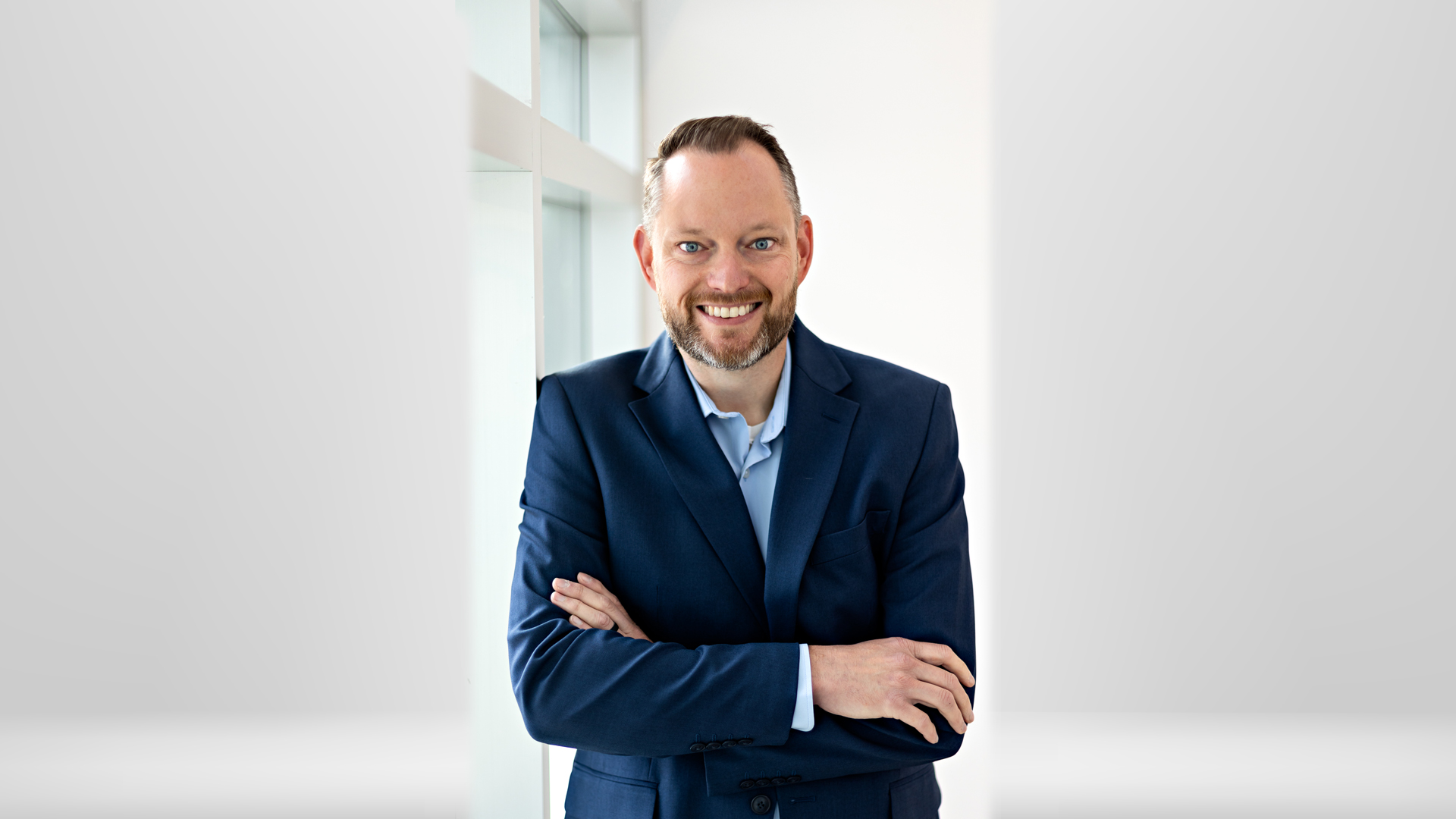 Director of TRIO Student Support Services Matt Stewart smiling beside a tall window in the Besse Gallery wearing a navy suit jacket and French blue shirt with his arms crossed.