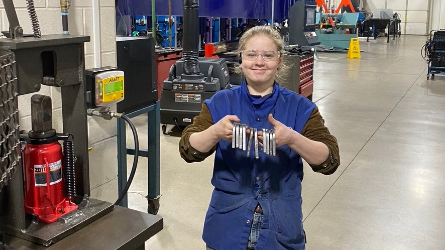 Recent welding graduate Olivia Trulock smiles in the welding lab wearing a welding apron and safety glasses while holding up sample welds.