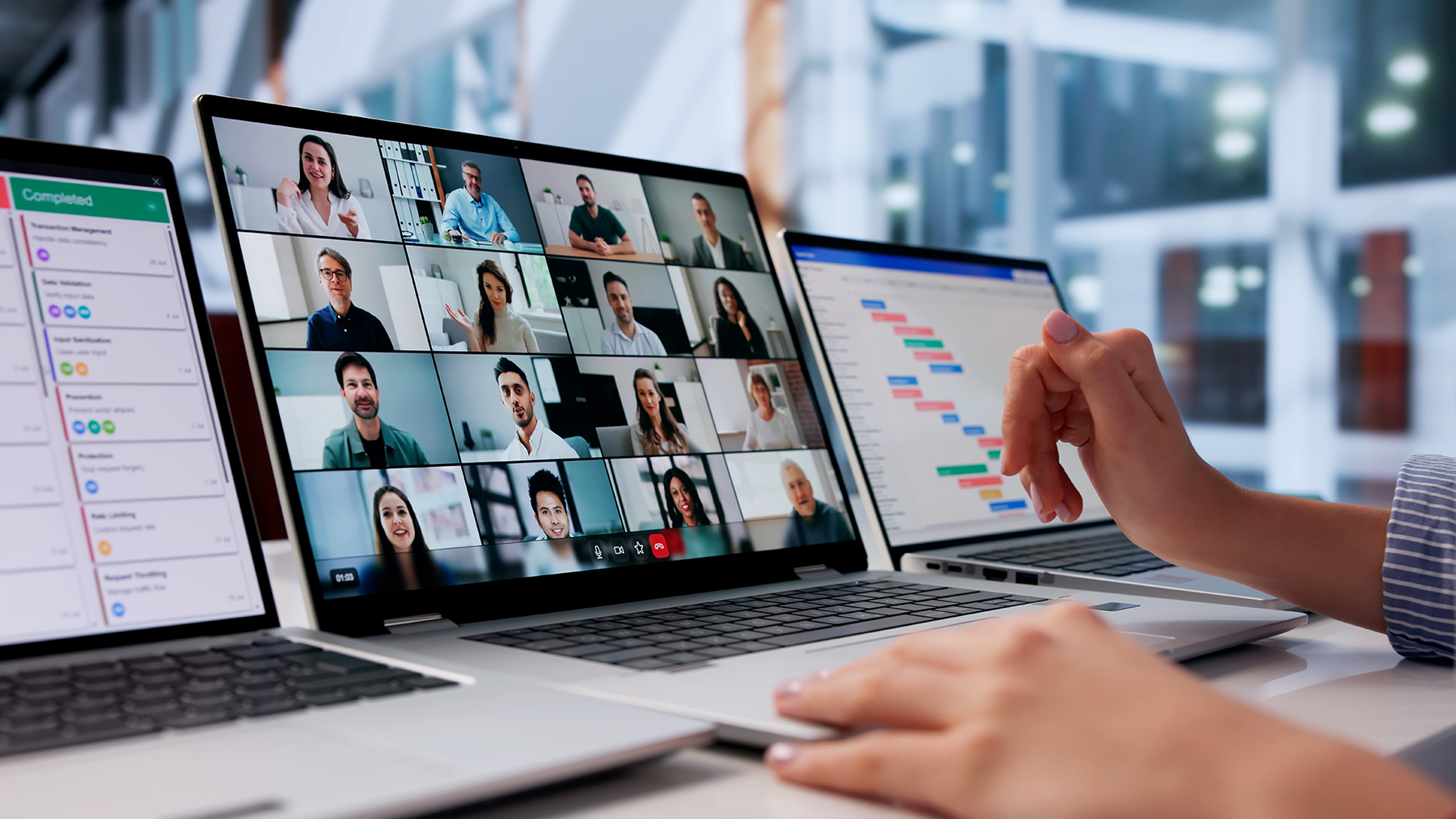 Stock image of a person sitting in on an online class, showing students on one screen, data on a second screen, and a gant chart on a third screen..